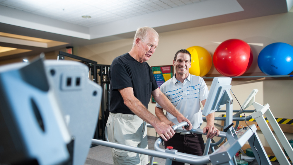 older man walking on treadmill next to physical therapist
